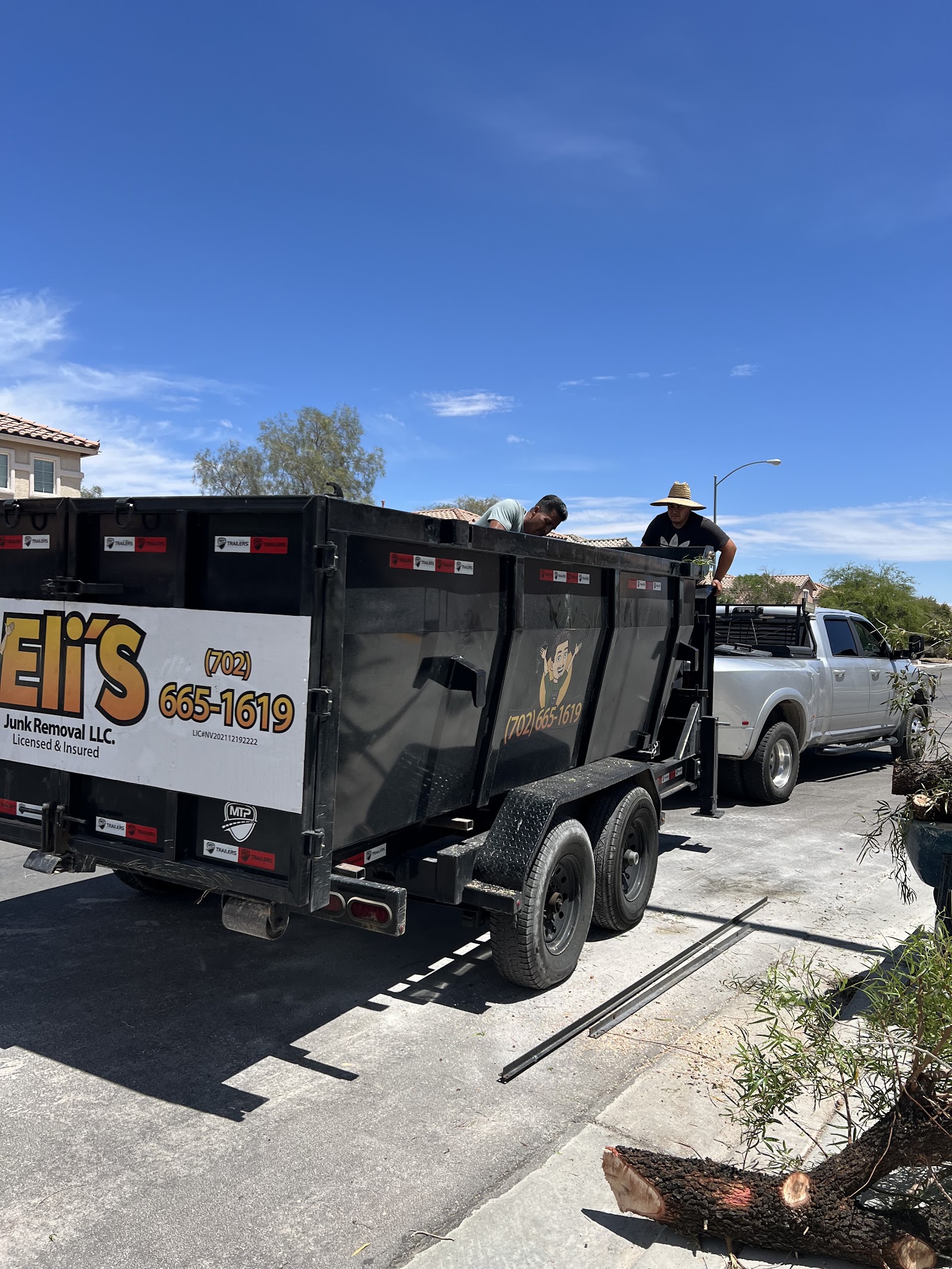 Eli's Junk Removal branded dump trailer and truck with crew loading tree debris in a residential neighborhood
